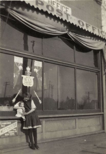 Nola Franklin (daughter of Jerome Francis Franklin Jr.) holding a baby doll. It is summer and she is standing in front of the Franklin Hardware Store and Eland Post Office, possibly next to the Franklin family Drug Store.