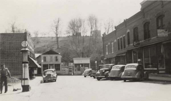 Street view of buildings in the center of a small town. Sidewalks run along both sides of the street. Automobiles are parked diagonally on the right and along the curb on the left. In the distance are more buildings on top of a hill behind trees. On the right is a sign for Potosi Beer, and a man is next to a gas pump on the left.