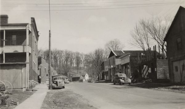 Street view of buildings in the center of a small town. A sidewalk runs along the left side of the street, and automobiles are parked on both sides. In the distance are more buildings at the base of a hill with trees on it. In 1938, East Main Street/Highway 133 became the Great River Road.