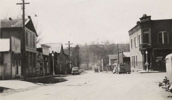 Street view of buildings in the center of a small town. Sidewalks run along both sides of the street and automobiles are parked along the curbs. On the corner on the right is a tavern. Date stone at the top of the building reads: "1900, W. Welsh," and in the window a sign reads: "Corner Beer." Over the door is a balcony with a "Potosi Beer" sign under it. On the left side of the street is a building called "Chicken Inn." In the distance are more buildings in front, and in the background behind trees is a hill. In 1938, East Main Street/Highway 133 became the Great River Road.