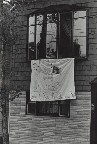 Hand-drawn banner hanging from the bay window of a home. The banner features a cartoon, rear view, of a boy holding an American flag, with his arm around a girl. They are standing in front of the door into South Boston High School and are not wearing any clothes. The text reads: "Love Is Going to Your Neighborhood School." The banner is most likely a protest against court-ordered busing to achieve integrated schools in Boston.