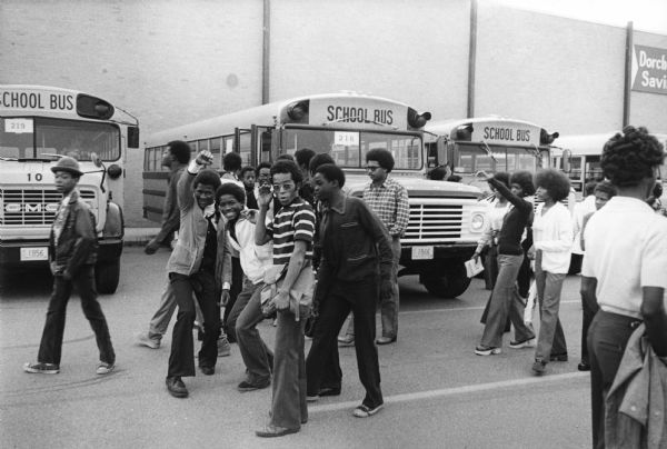 Students from Columbia Point waiting outdoors to be bused to South Boston High School. Most of the students are walking, and a few are clowning for the camera. School buses are parked in the background.