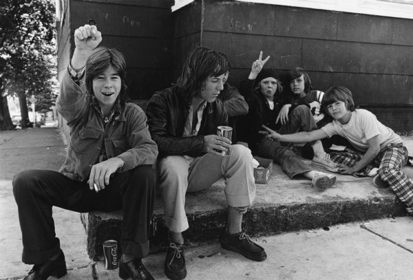 A school boy is raising a fist for the photographer while sitting with a group of his friends. Handwritten caption on the reverse: "These boys asked us to caption photo 'Tell them Southie hates niggers.' They are boycotting S. Boston High, looking for action."