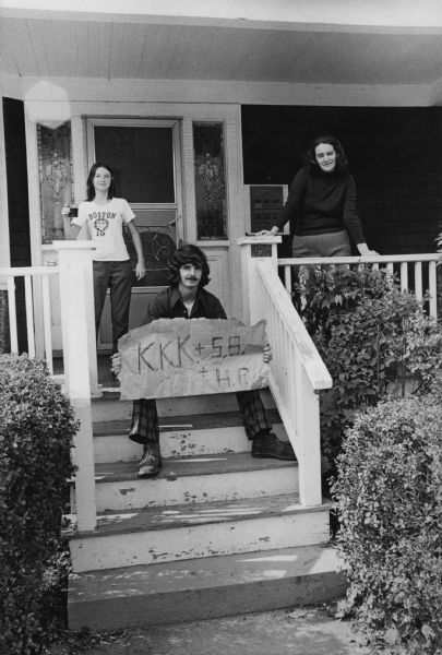 A man, sitting on porch steps, is holding up a sign that reads: "K.K.K. + S.B + H.P." (Ku Klux Klan plus South Boston plus Hyde Park.) Standing on the porch is a woman leaning on the railing on the right, and a teenage girl in a Boston t-shirt standing on the left and holding a beverage.