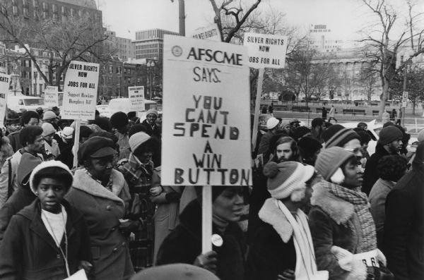 Demonstrators at a PUSH (People United to Serve Humanity) March supporting the Hawkins Humphrey Bill. Protest signs worn and held by the demonstrators read: "Silver Rights Now, Jobs for All, Support Hawkins-Humphrey Bill." The large sign in the center reads: "AFSCME Says, You Can't Spend A WIN Button." WIN stands for "Whip Inflation Now," a failed program started by President Gerald Ford. Caption on the reverse reads: "March around the White House 46th Anniv. of MLK birthday."