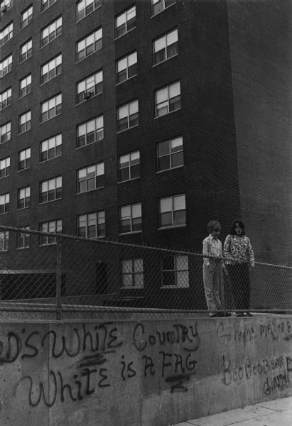 Two students are standing behind a fence enclosing the yard of a housing project. Below their feet is a concrete retaining wall with racist graffiti spray painted on it, which reads: "This is God's White Country, Kevin White is a Fag, Go Home Mayor Black, and BooBooBear Freshie." Kevin White was the the Mayor of Boston at that time. Caption on the reverse reads: "Two kids playing in project yard in South Boston, boycotting their school to protest busing."