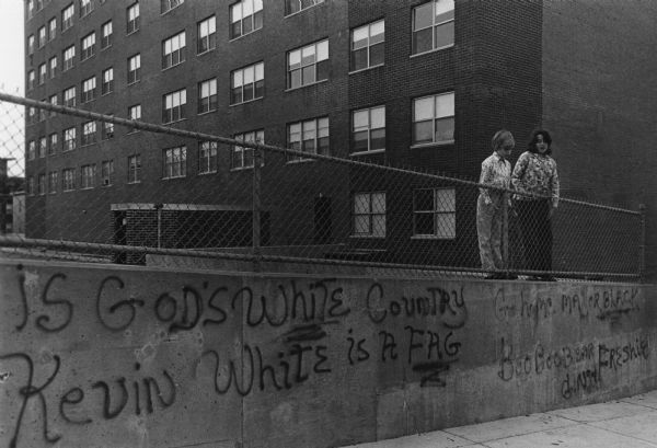 Two students stand next to a fence enclosing the yard of a housing project. Below their feet is a concrete retaining wall with racist graffiti spray painted on it, which reads: "[This] is God's White Country, Kevin White is a Fag, Go Home Mayor Black, and BooBooBear  Freshie." Kevin White was the the Mayor of Boston at that time. Caption on the reverse reads: "Two kids playing in project yard in housing project in South Boston, boycotting their school to protest busing."
