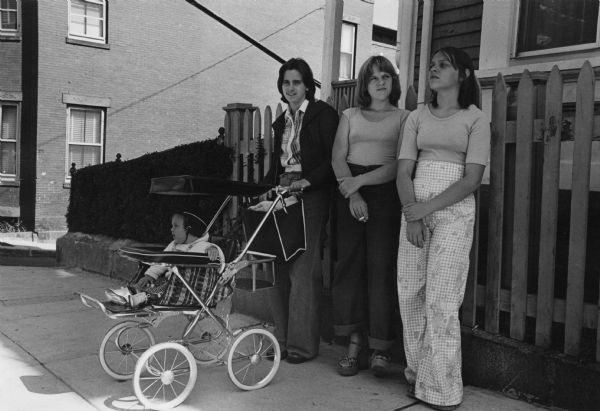 A recent graduate of South Boston High School and her friends are standing on the sidewalk in front of a house. One of the woman has a baby in a baby stroller. In the background is a brick building. This image was captured in South Boston during the unrest following court-ordered school desegregation busing.