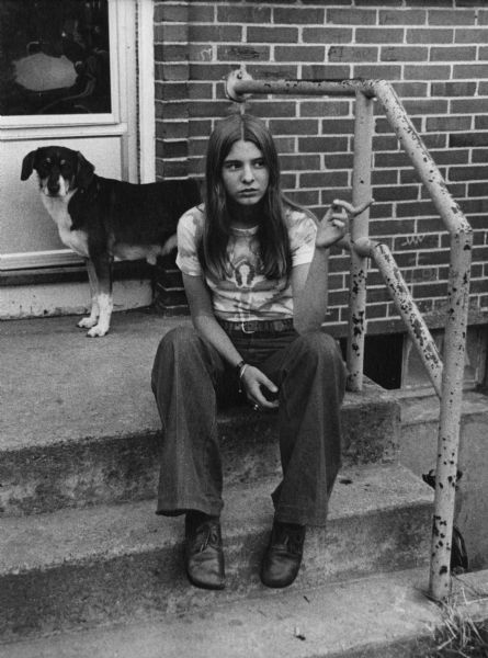 A teen girl is sitting on the steps leading up to a doorway, pointing to something on the right. A dog is standing behind her. This image was captured in South Boston during the unrest following court-ordered school desegregation busing.
