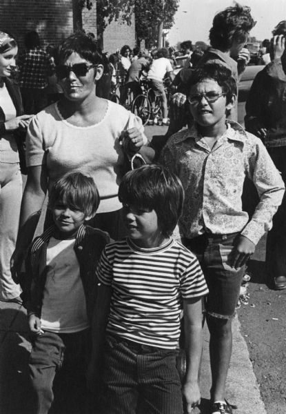 Donna Connolly, wearing sunglasses, is standing on the sidewalk in South Boston with some of her children. She was one of the leaders of the school boycotts to protest court-ordered busing in her city. Behind her are several other people, and a group of youth on bicycles are in the background.