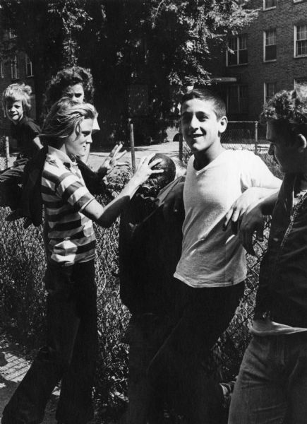 A group of students in South Boston are boycotting school to protest court-ordered desegregation busing. One boy is grabbing the head of an effigy of an African-American man. Two of the boys are sitting on a chain-link fence. In the background are trees and a housing project.