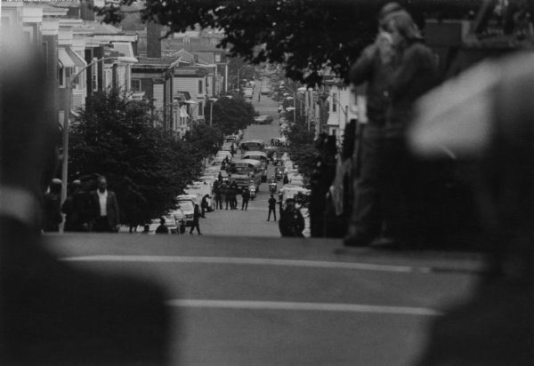 View down street towards school buses transporting minority students to South Boston High School. They were escorted by police. Protestors are lined up along the street, hurling insults and objects at the buses. Windows were broken and students were injured by flying glass.