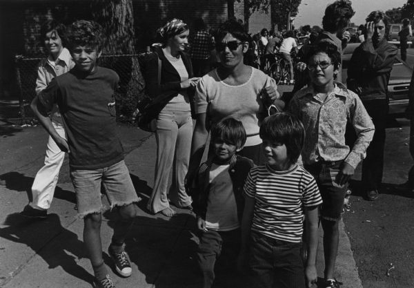 Donna Connolly, wearing sunglasses, is standing on the sidewalk in South Boston with some of her children. She was one of the leaders of the school boycotts to protest court-ordered busing in her city. Behind her are several other people, and a group of youth on bicycles are in the background. In the background on the right, standing in the street, is a police officer wearing a helmet. In the background are buildings and automobiles.
