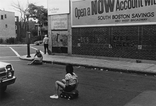 Two girls are rolling about in the street on small, foot-powered, six-wheeled ATV toys, and a man is standing on the corner next to a building with a billboard. Under the billboard is graffiti spray-painted on the brick wall that reads: "Kill Niggers." The street intersects with Dorchester Street, the route of the school buses that transported minority students to South Boston High School. This image was captured during the unrest following court-ordered school desegregation busing. 