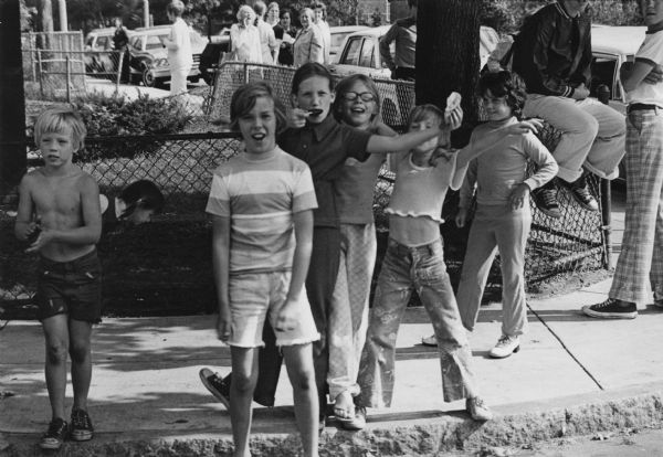 Students from South Boston are posing and clowning on a curb near a street corner. Older students are standing or sitting on chain-link fence. In the background, are adults on a sidewalk. This image was captured during the unrest following court-ordered school desegregation busing.