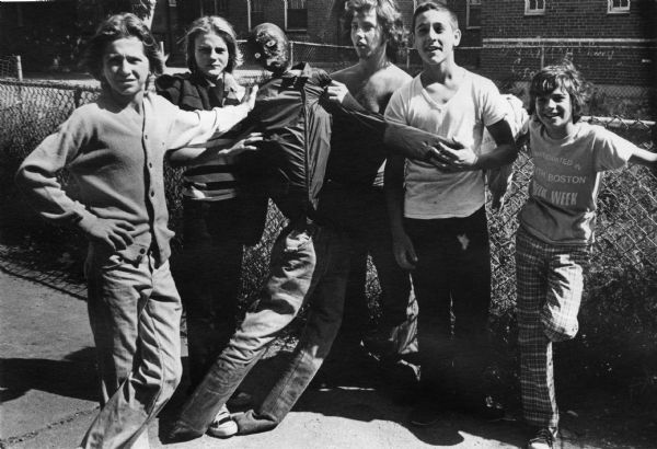 A group of South Boston teenagers posing with an effigy of an African-American man. In the background is a chain-link fence, shrubs and a housing project. This image was captured during the unrest following court-ordered school desegregation busing.