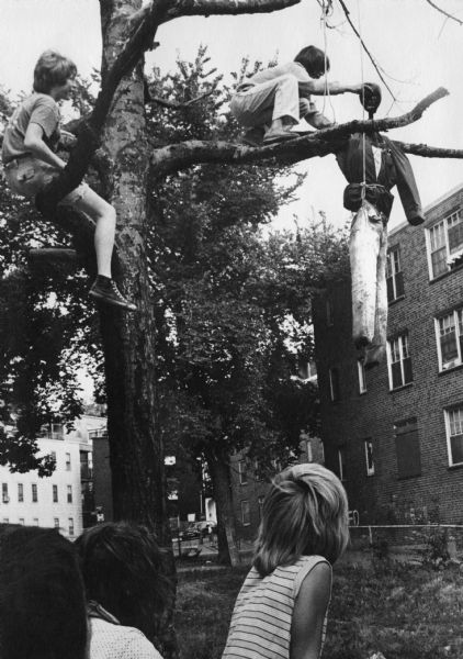 A group of children are hanging an effigy of an African-American man in a dead tree, creating a mock lynching. One of the boys is sitting on a limb of the tree, and another boy is arranging the effigy in the fork of a branch. Other children are watching in the foreground. In the background are shrubs, trees and a housing project. This image was captured during the unrest following court-ordered school desegregation busing.