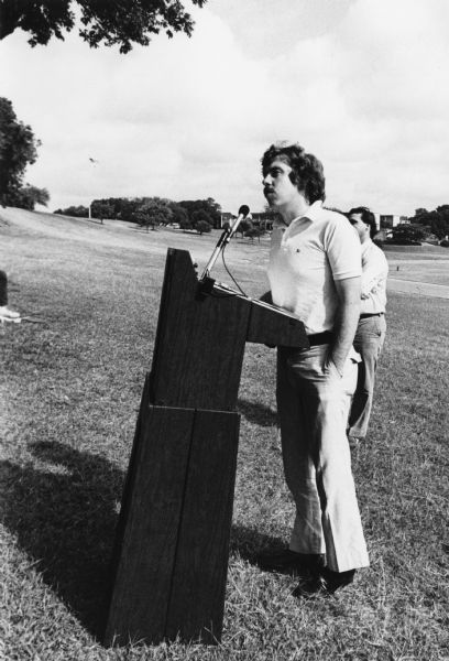 Activist Sam W. Brown, Jr., speaking while standing at a podium on a lawn in a large outdoor space. The event is the Conference on Alternative State and Local Public Policies held at the St. Edward's University Campus. Brown was the Colorado State Treasurer from 1975 to 1977.