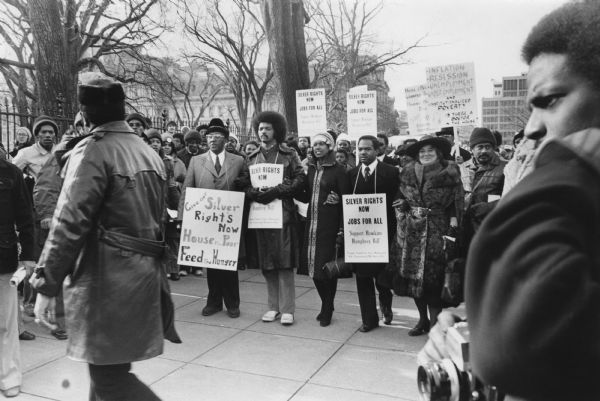 Demonstrators at a PUSH (People United to Serve Humanity) March walking around the White House to commemorate the 46th anniversary of the birth of Martin Luther King Jr. In the center, (left to right), are an unknown man, the Reverend Jesse Jackson, unknown woman, Delegate Walter E. Fauntroy and Representative Bella Abzug. The march also supported the proposed Humphrey-Hawkins Full Employment Bill. An estimated 2000 people participated, carrying signs. Some of the signs that people are carrying read: "Silver Rights Now, Jobs for All, Support Hawkins-Humphrey Bill," Give Our Silver Rights Now, House the Poor, Feed the Hungry," and "Inflation, Recession, Unemployment, Under-Employment and Institutionalized Poverty, Is There a Doctor in the House?" A man standing in the foreground on the right is holding a camera.