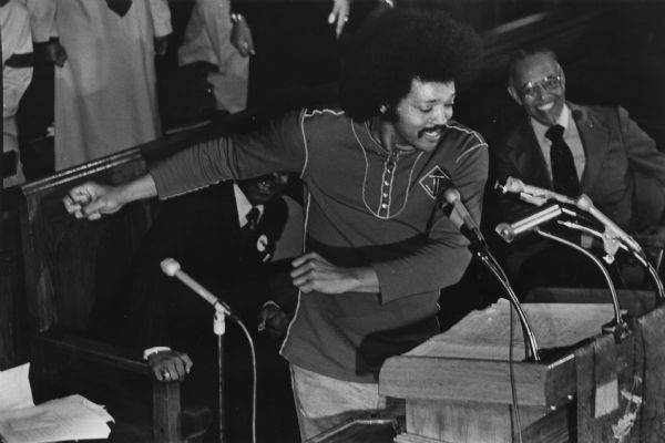 The Reverend Jesse Jackson preaching at a pulpit with microphones at Shiloh Baptist Church. Members of the congregation, and singers in robes can be seen behind him. On the right a man is smiling broadly.
