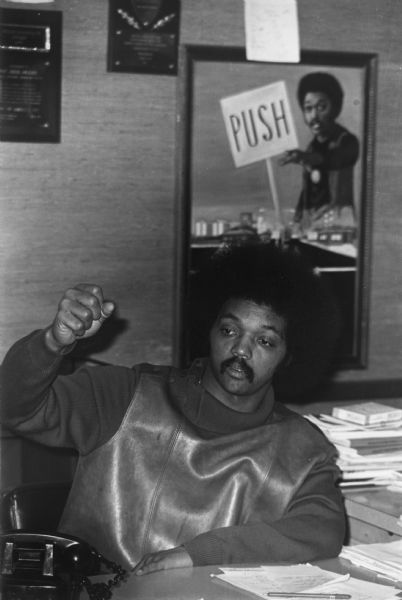 The Reverend Jesse Jackson gesturing while sitting at his desk in the PUSH (People United to Serve Humanity) headquarters. In the background, on the wall, is a painting of Jackson with a PUSH sign, and two awards.