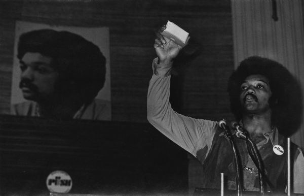 The Reverend Jesse Jackson addressing an Operation PUSH (People United to Serve Humanity) rally in its headquarters. He is wearing a PUSH button on his leather vest, and he is holding a small box in his uplifted hand while he is speaking. On the back wall is a large portrait of Jackson.