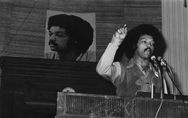 The Reverend Jesse Jackson addressing an Operation PUSH (People United to Serve Humanity) rally in its headquarters. He is wearing a PUSH button on his leather vest, and is pointing with one finger while speaking. On the back wall is a large portrait of Jackson.