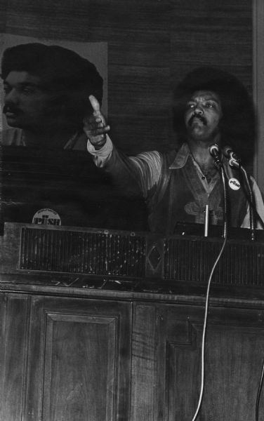 The Reverend Jesse Jackson addressing an Operation PUSH (People United to Serve Humanity) rally in its headquarters.  He is wearing a PUSH button on his leather vest, and is gesturing with one hand while speaking. On the back wall is a large portrait of Jackson.