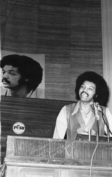 The Reverend Jesse Jackson addressing an Operation PUSH (People United to Serve Humanity) rally in its headquarters. He is wearing a PUSH button on his leather vest. On the back wall is a large portrait of Jackson.