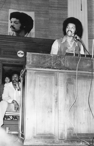 The Reverend Jesse Jackson addressing an Operation PUSH (People United to Serve Humanity) rally in its headquarters. On the back wall is a large portrait of Jackson. He is wearing a PUSH button on his leather vest. A man and woman are sitting behind the podium on the left.