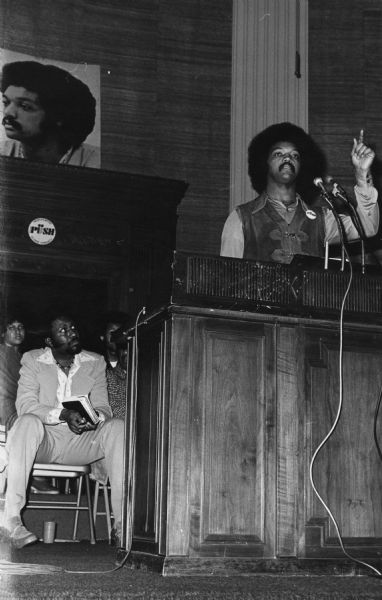 The Reverend Jesse Jackson addressing an Operation PUSH (People United to Serve Humanity) rally in its headquarters. He is pointing with one finger while speaking. On the back wall is a large portrait of Jackson. He is wearing a PUSH button on his leather vest. Three people are sitting in chairs behind the podium on the left.