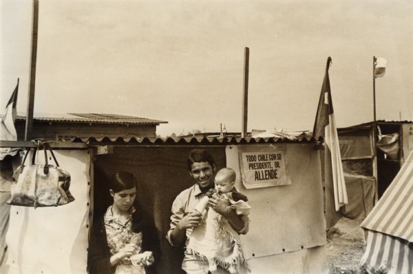A family of squatters standing in front of a shack, in a camp outside of Santiago, Chile. The father is holding a child in his arms and the mother is standing on the left. A duffel bag is hanging on a beam, and Chilean flags are flying over the camp. The sign on the wall reads: "Todo Chile con su Presidente, Dr. Allende" translates as: "All Chile with its President, Dr. Allende."