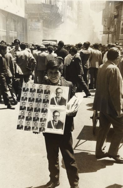 A smiling young man is selling pictures of President Allende on the street on Inauguration Day. He was the first Socialist President of Chile. In the background a crowd is in the street, and dust or smoke is rising between buildings.