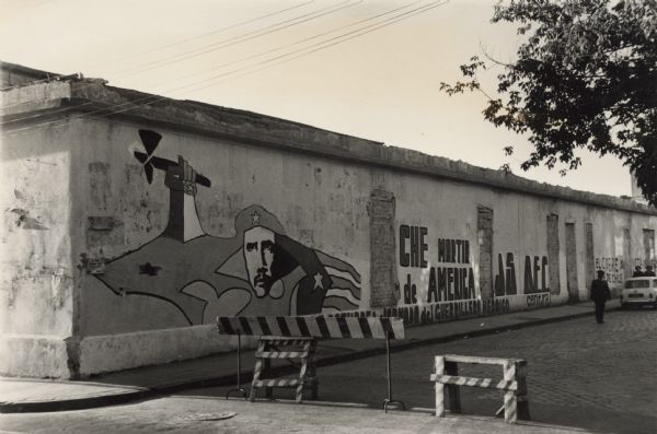 View from intersection towards a blocked off street with political graffiti on the wall of a building in Santiago, Chile. On the left is a a depiction of Che Guevara, a flag and a hand holding an axe. Further down the street on the right are pedestrians and an automobile parked along the curb. Caption written on the back reads: "'Che Martir de America,' and  '[???] Day of the Heroic Guerrilla.'"