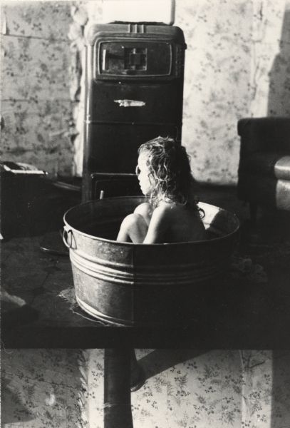A young girl is sitting in a galvanized washtub on the floor in front of an oil burning stove and an armchair. Flowered wallpaper covers the walls. [The bottom of the print includes the top of the adjoining negative.]