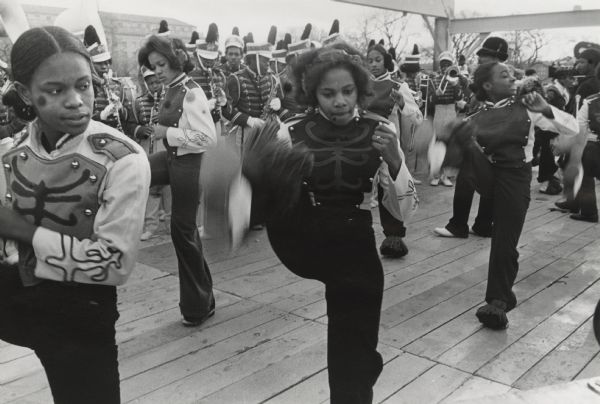 Dancers in uniform performing in the foreground, with a marching band in the background. Pompoms are attached above the dancers shoes. They are performing in an open structure with a wooden floor. In the background on the left is a large building.