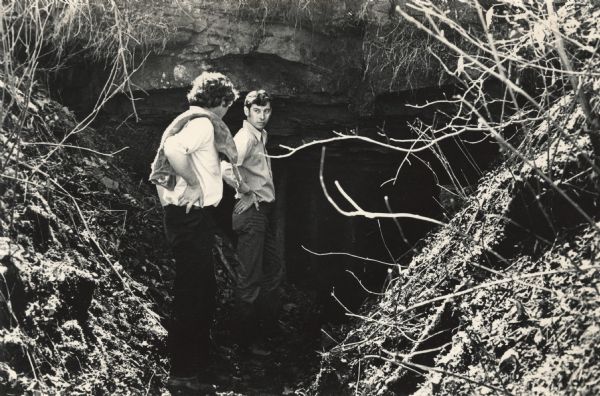 Two men are standing in front of the entrance to a mine. Vertical beams are just inside the entrance. The man on the left has a cloth sack draped over his shoulder.