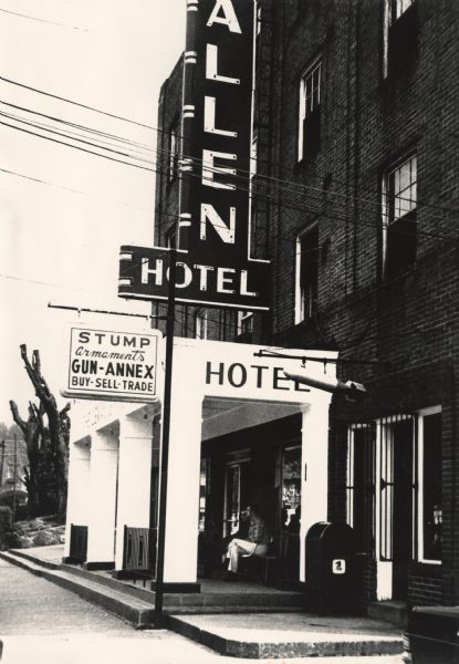 Exterior view of the entrance to a hotel and a gun store. Two people are sitting under the portico. A replica of a bomb is hanging from a pole near the entrance over a post office box The tall, neon hotel sign reads: "[Lew]allen Hotel." The gun store sign reads: "Stump Armaments, Gun-Annex, Buy-Sell-Trade." 