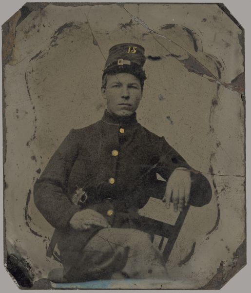 Ferrotype/tintype of unknown soldier. Three-quarter length portrait, facing forward, sitting in a chair sideways with his legs crossed. He is in uniform with a cap and sword. Buttons, buckle on cap and the number 15 on his cap have been highlighted with gold details. The outline of a nonpareil mat is on the tintype.