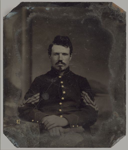 Ferrotype/tintype of unknown soldier in front of a painted backdrop. Three-quarter length portrait, facing forward, sitting in a chair with his legs crossed. He is in uniform with 3 bar Sargent's chevrons on the sleeves, and his buttons have been highlighted with gold details. His cap is on the table next to his elbow. The outline of a nonpareil mat is on the tintype.