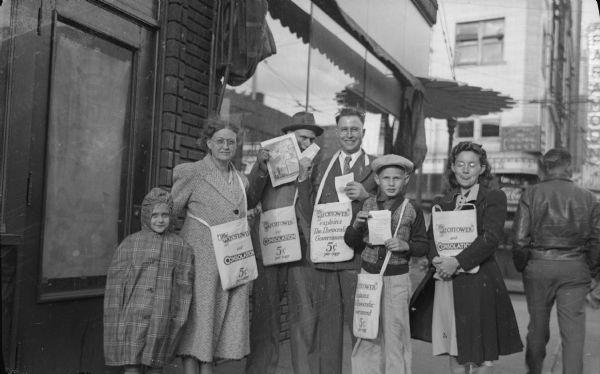 A group of Jehovah's Witnesses selling copies of <i>The Watchtower</i> magazine. Standing together on a sidewalk are two women, two men, a girl and a boy. They are each wearing a bag slung over their shoulders, except for the girl on the far left. Text on the bags reads: "The Watchtower and Consolation. 5¢ per copy," and "The Watchtower explains The Theocratic Government. 5¢ per copy." The men and the boy are displaying literature.