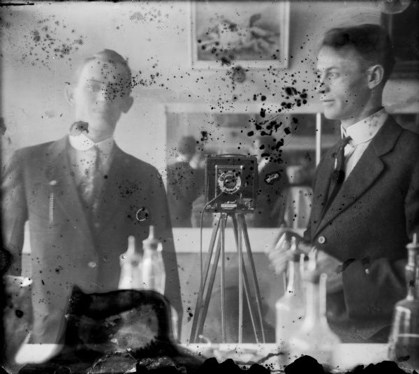Double-exposed, self-portrait of the photographer looking into a mirror, with another mirror in the background. He is wearing a suit, and has changed a few accessories between exposures. The camera is in the middle of the image. Bottles are on the counter in the foreground.