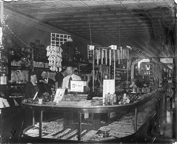 Interior of a Dry Goods or a 5 & 10 Cent Store. Two men and a woman are standing on the left side of the large central display counter. Some of the products on display in the glass case include: handkerchiefs, jewelry, buttons, handbags, clothing, celluloid figures, papier-mâché Halloween decorations, Colgate Dental Ribbon Cream, toothbrushes, talcum powder and hair brushes. The ceiling is of pressed tin.