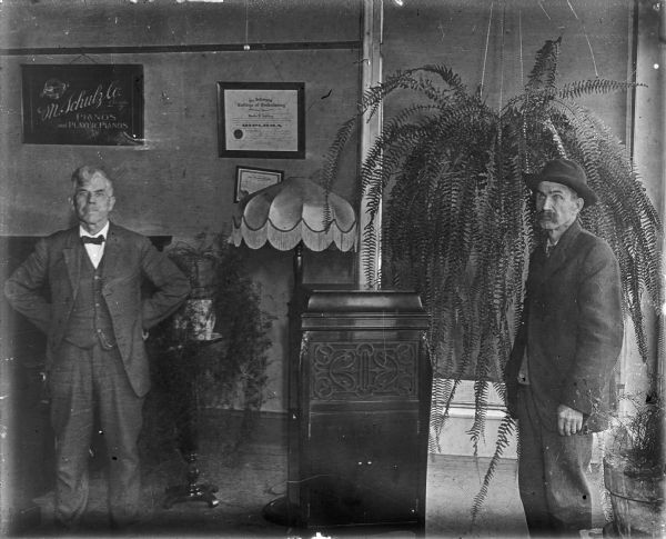 Two men are standing indoors, one man on the left and the other on the right. In the center is a vintage hand crank phonograph. A sign hanging on the back wall reads: "M. Schulz Co. Chicago, Pianos and Player Pianos." A Diploma reads, "The Johnson College of Embalming." A piano, large hanging Boston fern, asparagus fern on a stand and a floor lamp with a fringed lampshade are in the background.