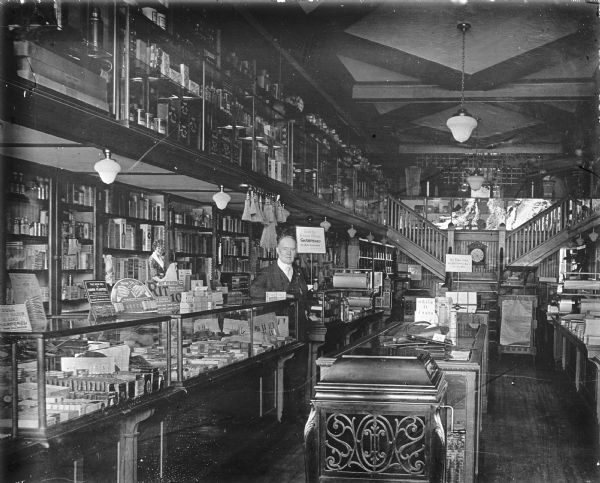 A man stands between display cases in a general store, behind him on shelves are many bottles in boxes and wrappers. On the counters are displays for Rit Dye, corn plasters, New Skin, razor blade sharpening and Parker Pens. Whisk brooms hang from the ceiling. A vintage hand crank phonograph is in the foreground. The mezzanine has more counters and shelving. The store is illuminated with electrical lights.