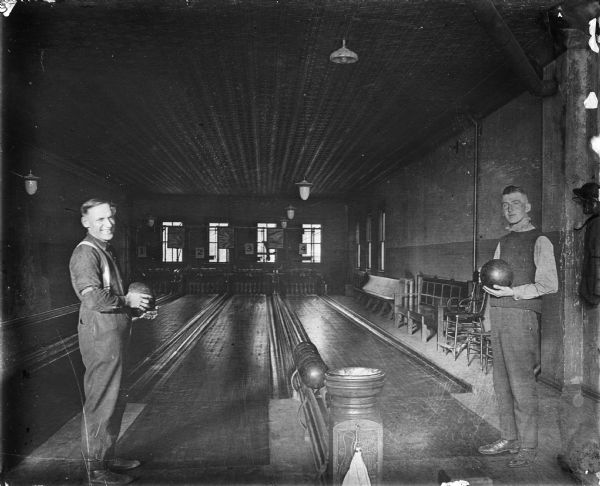 Two men stand in a four lane bowling alley. Each man is holding a bowling ball. Between them is a Brunswick Balke Collender ball return post with a towel hanging on a string from a hook. All four lanes feed into one ball return. On the right is seating for spectators. The ceiling is made of pressed tin.