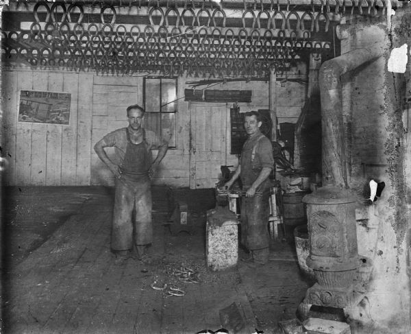 Two men are standing in a blacksmith shop. One man is working at an anvil, the other stands with his hands on his hips. They are wearing leather aprons. Hundreds of horseshoes hang from nails in the ceiling joists. There is a small pile of horseshoes on floor with metal fragments. On the right is an ACME Oaklet pot belly stove made in Newark, Ohio by the Wehrle Company.