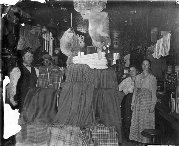 Group portrait of people posing in a dry goods shop. Two men are on the left, and two women are on the right. Cloth fabric, solids and plaids, are on display in the foreground, and pelts are hanging from the ceiling. Clothing is hanging on display along the wall on the right. A "Selz Shoes" sign hangs on the back wall.