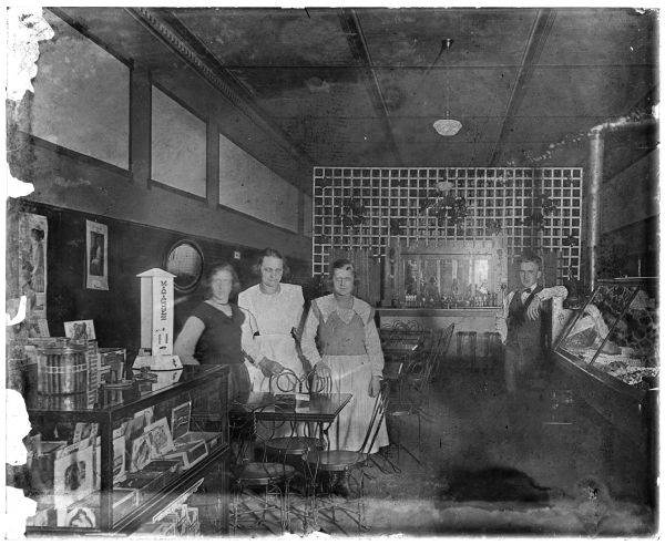 Group portrait of people standing and posing in a restaurant/soda fountain. Three women are standing on the left near a twisted wire table and chairs, a glass case displaying cigars and a large Northwestern Corp Porcelain 1 Cent Match dispenser. Above are Cola Cola calendars and an oval mirror. A man is leaning on a cash register near a glass display case of candy on the right. There is a mirror surrounded by a lattice along the wall in the background, perhaps a soda fountain. 
