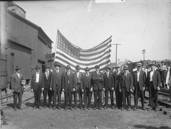 Group portrait of men lined up outdoors in a train yard, in front of an American flag. They are all wearing suits and hats, and three men have boutonnieres. One man is smoking a cigar and the man on the far left has two cigar boxes under his arm. Two women stand behind the men, holding the flag and other people are in the background. One boxcar has "Chicago, Milwaukee and St. Paul" on the side. Depot buildings and rail cars can be seen all around.